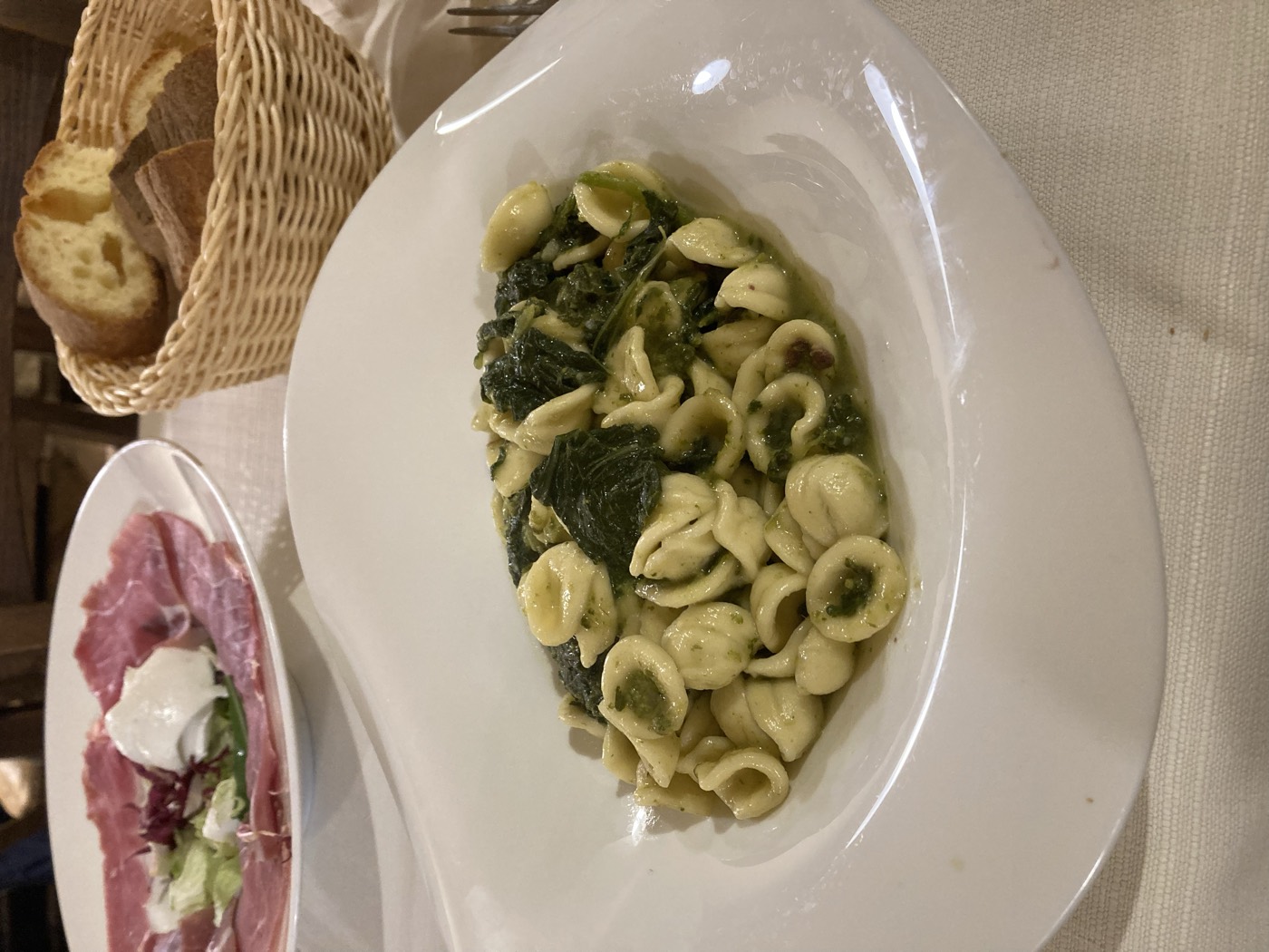 a plate of orecchiette with greens, with cured ham and a bread basket behind